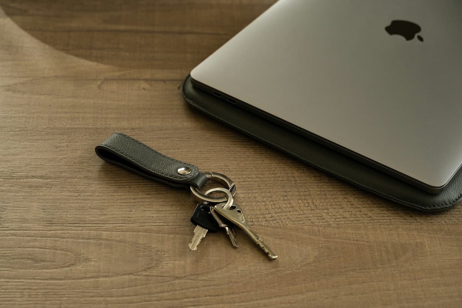 A closed laptop and a set of keys on a leather fob, resting on a wooden desk.