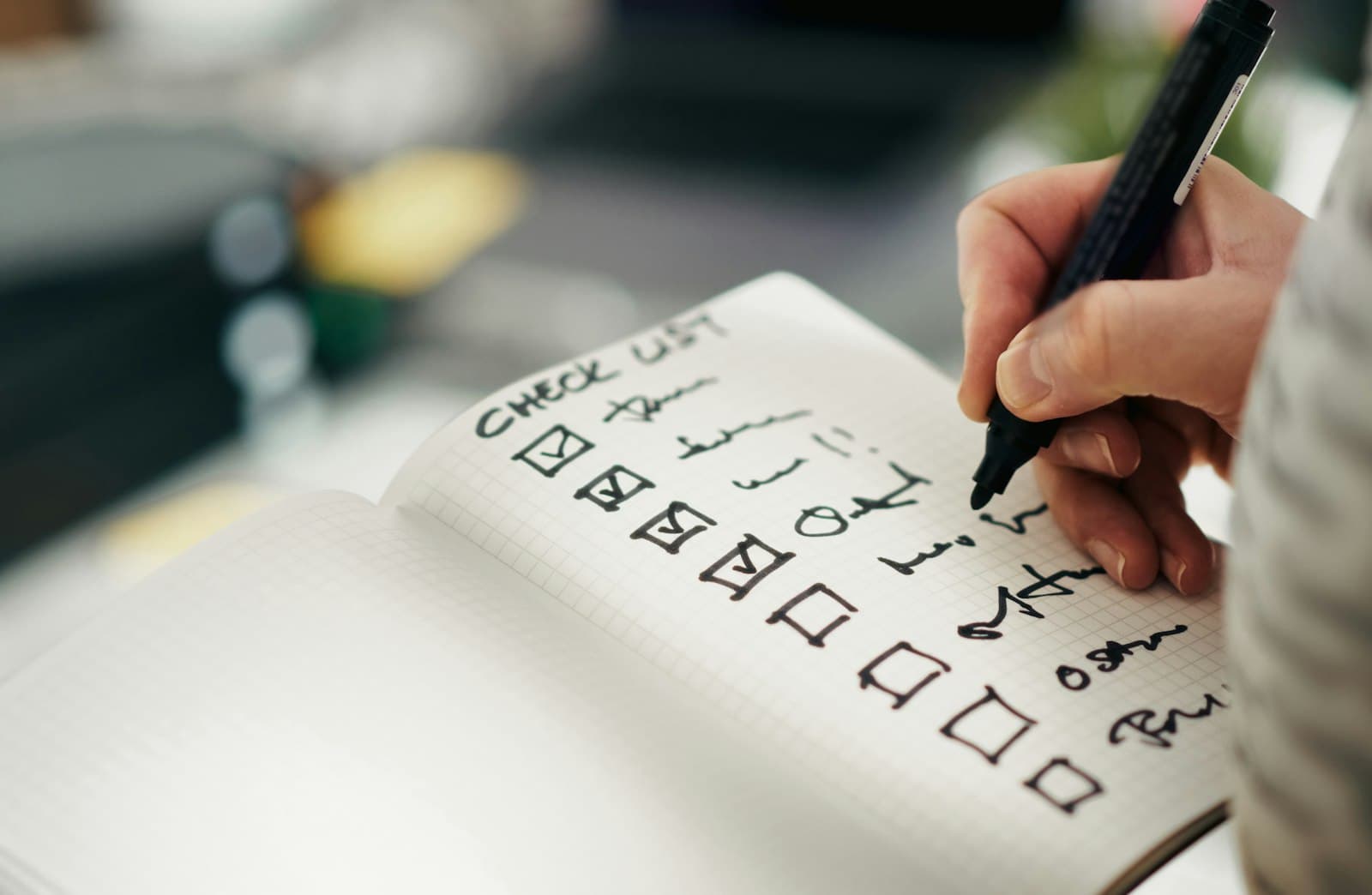 A hand ticking boxes on a handwritten checklist in a grid notebook.