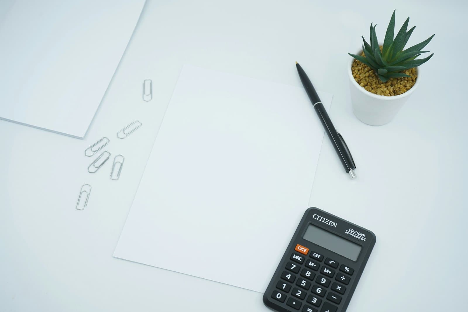 A calculator, pen and blank paper laid out on a white desk.