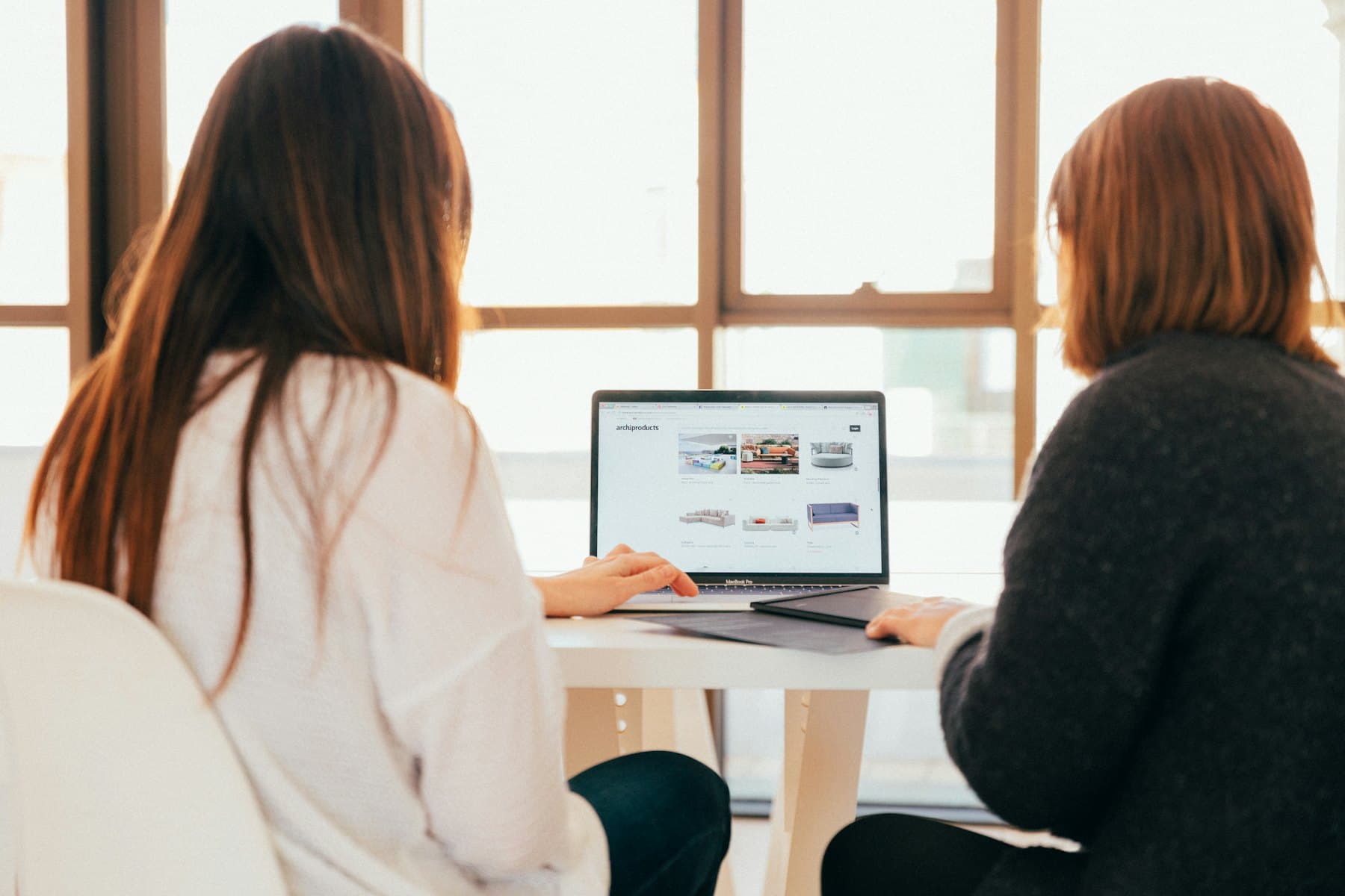 Two colleagues sitting side by side at a desk, looking at the same laptop screen.