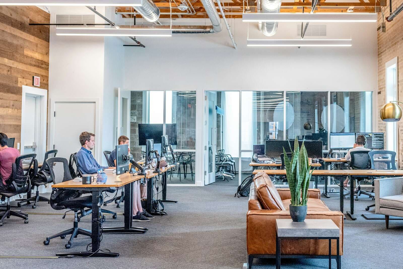 A bright open-plan office with exposed ceiling beams, a few people working at standing desks with monitors, and a leather sofa and potted plant in the lounge area.