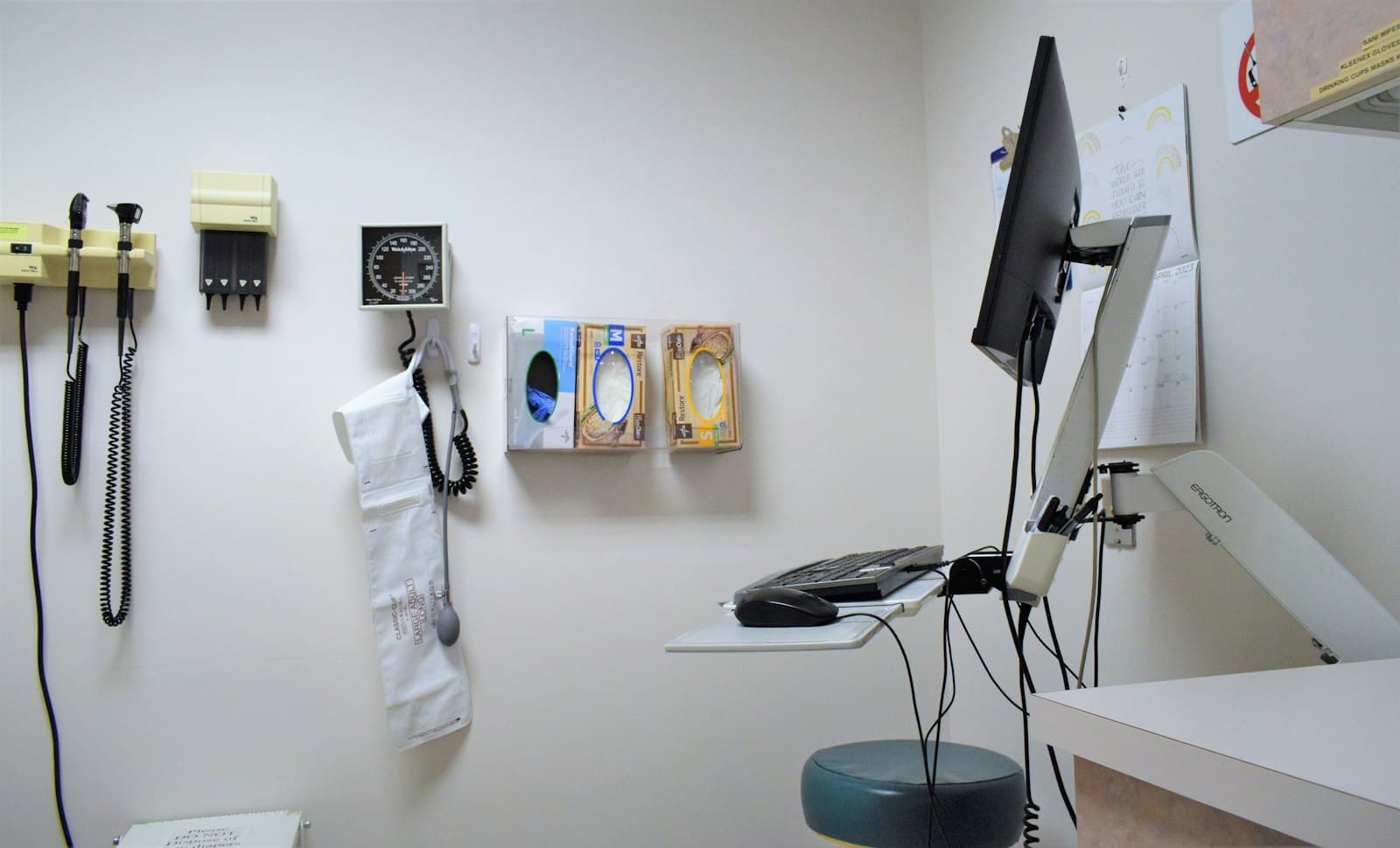 An empty exam room: wall-mounted otoscope and blood-pressure gauge, glove dispensers, and a computer workstation on an articulating arm.