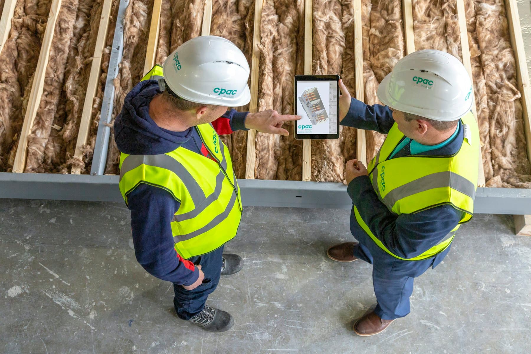 Two workers in hard hats and hi-vis vests looking at plans on a tablet inside a building under construction.