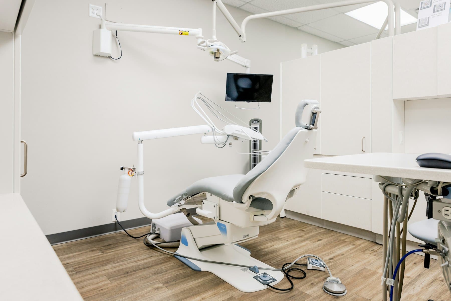 An empty dental treatment room with a chair, overhead light and a wall-mounted monitor.