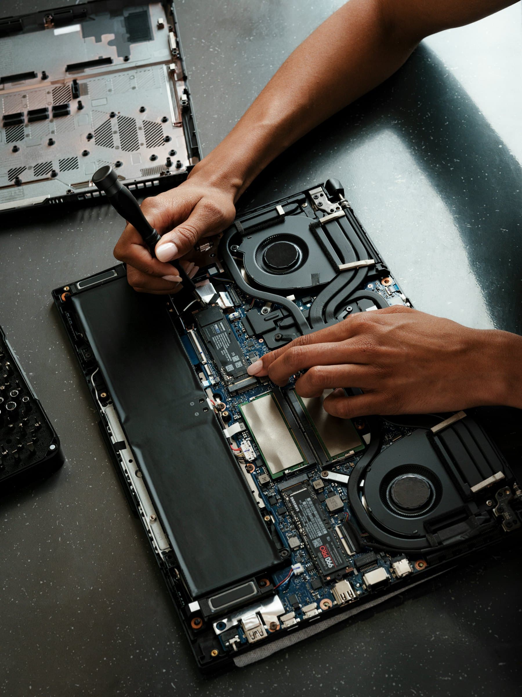 A laptop opened on a workbench, its battery and cooling fans exposed, while someone works on it with a screwdriver.